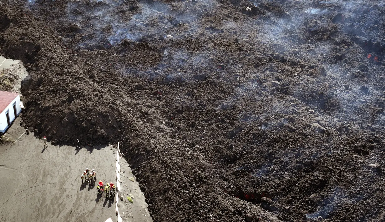 Petugas pemadam kebakaran berdiri di samping lahar yang mengalir saat gunung berapi terus meletus di pulau Canary La Palma, Spanyol, Senin (29/11/2021). Punggung bukit dan mengancam akan memperluas dampak pada tanah yang dievakuasi, infrastruktur dan rumah. (AP/Emilio Morenatti)