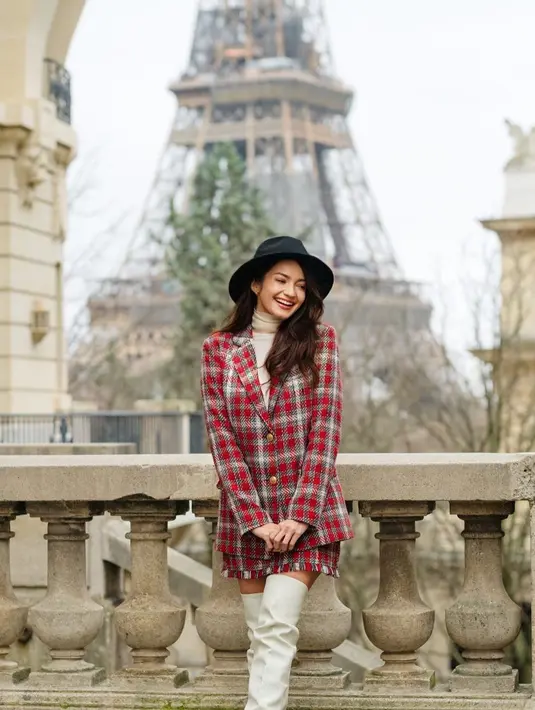 Enzy Storia berpose menawan di depan Menara Eiffel. Ia mengenakan set blazer dan roknya yang bermotif kotak-kotak merah. Enzy memadukan penampilannya dengan turtleneck cokelat, high boots putih, dan topi hitam. [Foto: Instagram/enzystoria]