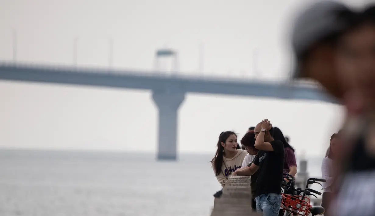 Orang-orang berdiri di pinggir pantai menghadap Jembatan Hong Kong-Zhuhai-Makau (HKZM) di Zhuhai (22/10). Jembatan laut terpanjang di dunia yang menghubungkan Hong Kong, Makau, dan daratan Cina ini akan dibuka 24 Oktober 2018. (AFP Photo/Fred Dufour)