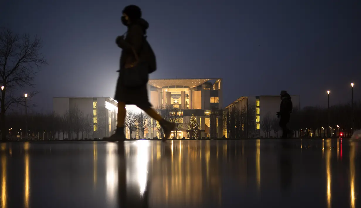 Seorang wanita berjalan di depan kanselir yang diterangi di Berlin, Jerman, Rabu (29/12/2021). Menteri Kesehatan Jerman Karl Lauterbach menyarankan warga Jerman untuk menghabiskan periode Tahun Baru 'sangat hati-hati' dan merayakan hanya dalam kelompok yang sangat kecil. (AP Photo/Markus Schreiber)