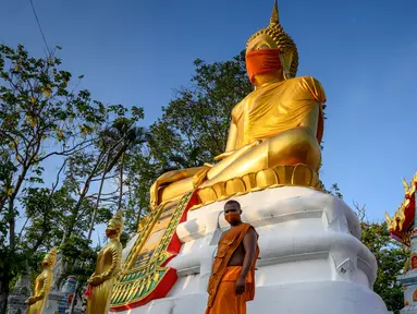 Biksu berjalan di depan patung Buddha raksasa yang mengenakan masker di kuil Wat Nithet Rat Pradit di Pathum Thani di luar Bangkok, Thailand, 12 Mei 2020. Pemasangan masker tersebut sebagai tanggapan terhadap penyebaran pandemi Covid-19. (Photo by Mladen ANTONOV / AFP)