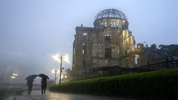 Dua pria berjalan di tengah hujan lebat di dekat Kubah Bom Atom di Hiroshima, Jepang barat,  Kamis (15/7/2021). Gedung ini dibiarkan begitu saja untuk memperlihatkan efek bom atom. Gedung ini berjarak sekitar 160 meter dari pusat ledakan (hypocenter). (AP Photo/Eugene Hoshiko)