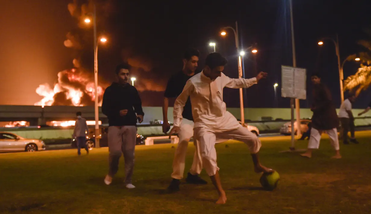 Anak-anak muda bermain sepak bola saat asap dan api membubung dari fasilitas minyak Aramco pada kejauhan menyusul laporan serangan pemberontak Yaman di Kota Jeddah, Arab Saudi, 25 Maret 2022. Pemberontak Yaman mengatakan mereka menyerang fasilitas minyak Arab Saudi tersebut. (AFP)