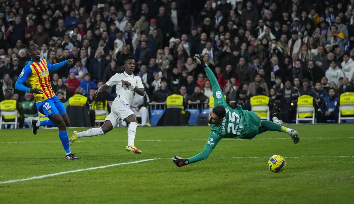 Penyerang Real Madrid, Vinicius Junior (tengah) menembak bola untuk mencetak gol melewati kiper Valencia Giorgi Mamardashvili selama pertandingan La Liga Spanyol di stadion Santiago Bernabeu di Madrid, Jumat (3/2/2023). Real Madrid menang atas Valencia 2-0 . (AP/Bernat Armangue)