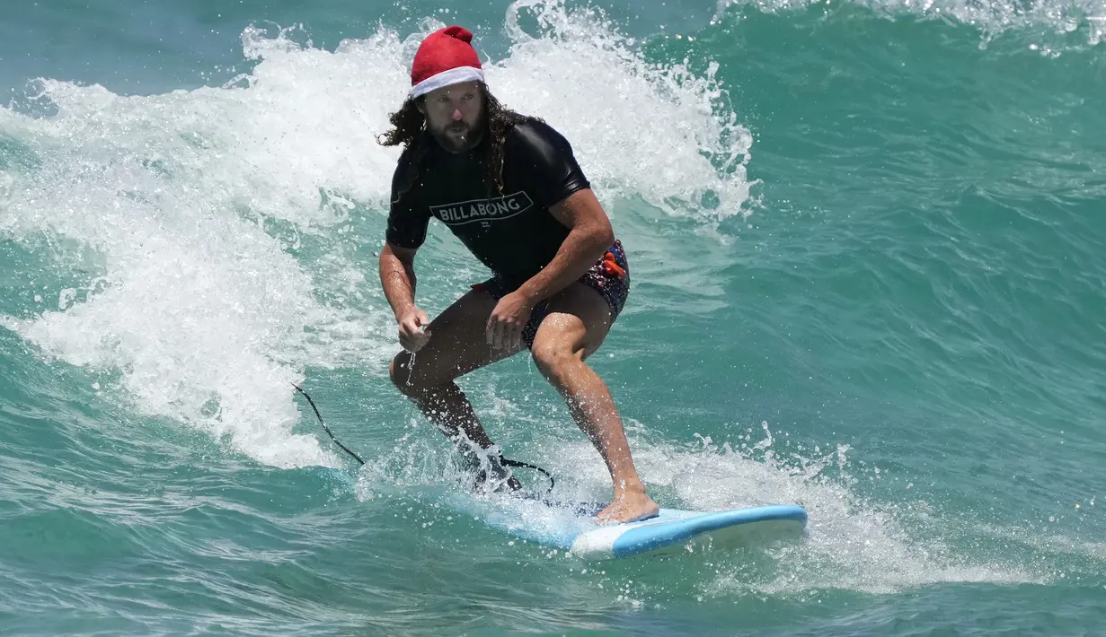 Tony O'Connor, asal Manchester, Inggris, naik papan selancar sambil mengenakan topi Santa saat merayakan Natal di Bondi Beach di Sydney, Sabtu (25/12/2021). (AP Photo/Rick Rycroft)