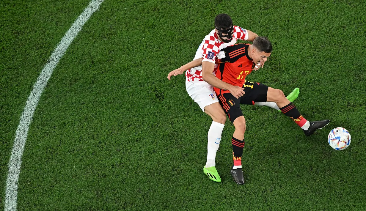 Bek Kroasia #20 Josko Gvardiol berebut bola dengan penyerang Belgia #17 Leandro Trossard pada laga terakhir Grup F Piala Dunia 2022 di Stadion Ahmad bin Ali, Kamis (1/12/2022). Josko Gvardiol yang memakai topeng pelindung wajah karena sempat mengalami cedera tersebut tampil impresif di laga Kroasia vs  Belgia. (MANAN VATSYAYANA / AFP)