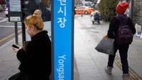 Seorang wanita bermain ponsel saat menunggu di halte bus di Seoul, Korea Selatan (19/2). (AFP Photo/David Gannon)