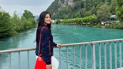 Titi Kamal saat berpose di pinggir sungai di Swiss. Titi tampil cantik dengan rambut terurai dengan senyum yang bahagia dengan mengenakan Sweater biru dan membawa tas berwarna merah saat menikmati keindahan sungai tersebut. (instagram/titi_kamall)