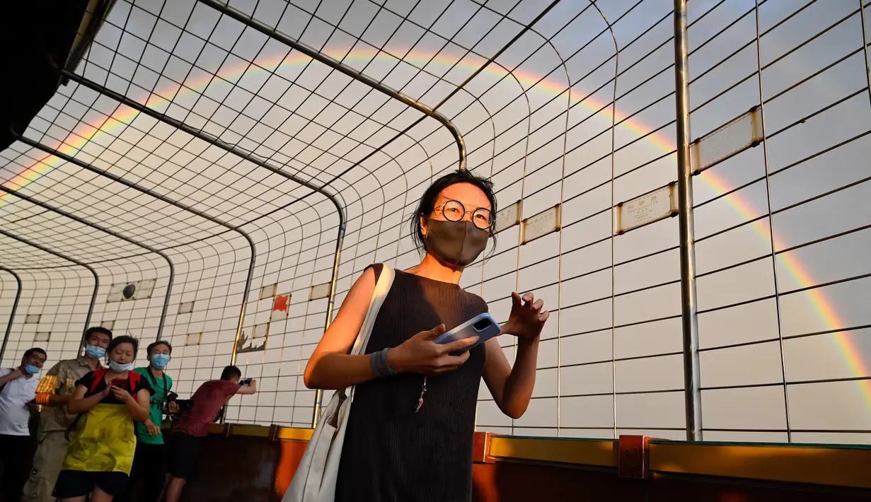 Pelangi terlihat melalui pagar saat seorang wanita berjalan di Central Television Tower di Beijing (26/8/2021). Keindahan pelangi ganda dimanfaatkan warga Beijing untuk diabadikan lewat foto dan berfoto dengan latar pelangi itu. (AFP/Jade Gao)