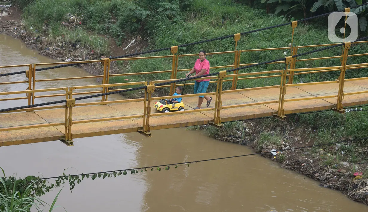 Seorang ibu bersama anaknya melintasi jembatan gantung Kuning yang menghubungkan Pasar Minggu Poltangan ke Cililitan Condet, Jakarta, Selasa (28/12/2021). Jembatan gantung Kuning tersebut merupakan jalan umum dan akses jalan warga yang ingin berlalu-lalang. (merdeka.com/Imam Buhori)