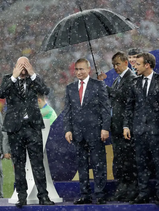 Presiden Rusia, Vladimir Putin dilindungi payung sementara Presiden FIFA Gianni Infantino dan Presiden Prancis Emmanuel Macron basah kuyup saat hujan turun selama penyerahan trofi Piala Dunia 2018 di Stadion Luzhniki, Minggu (15/7). (AP/Matthias Schrader)