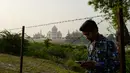 Seorang pria memainkan ponselnya di seberang Taj Mahal dari tepi sungai Yamuna di Agra, Jumat (4/5). Mahkamah Agung India meminta pemerintah mencari cara untuk mengembalikan warna asli situs bersejarah dunia, Taj Mahal. (AFP/CHANDAN KHANNA)