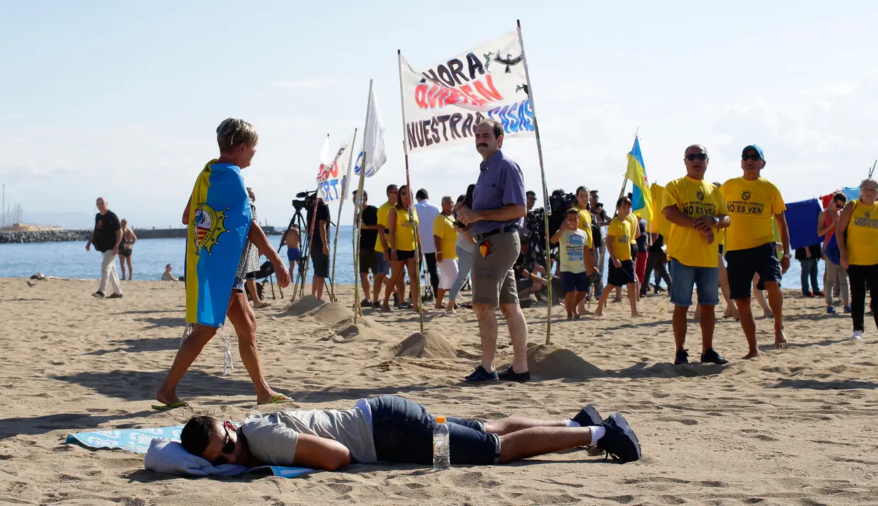 Seorang pria berbaring di atas pasir saat sejumlah orang melakukan aksi protes di pantai Barcelona, Sabtu (12/8). Para aktivis memprotes tempat-tempat populer di Spanyol melawan ledakan industri pariwisata yang tak terkendali. (AP Photo/Manu Fernandez)