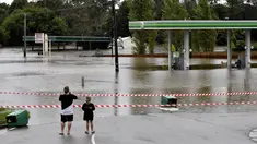 Sebuah keluarga berjalan mengunjungi pompa bensin yang terendam banjir di pinggiran barat daya Camden, Sydney, Australia, Selasa (8/3/2022). (Muhammad FAROOQ/AFP)