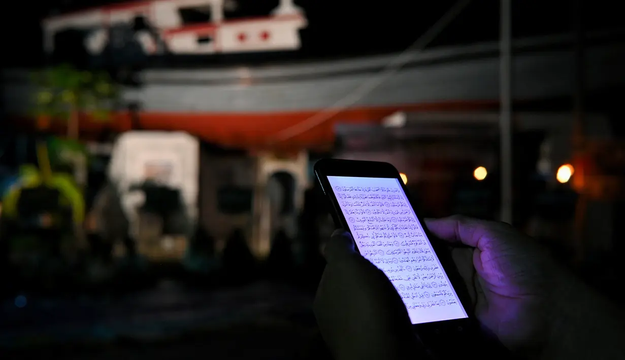 Seorang pria membaca Alquran di ponselnya saat upacara doa pada malam peringatan 17 tahun tsunami Boxing Day 2004 di monumen Kapal Tsunami Lampulo di Banda Aceh (25/12/2021). (AFP/Chaideer Mahyuddin)