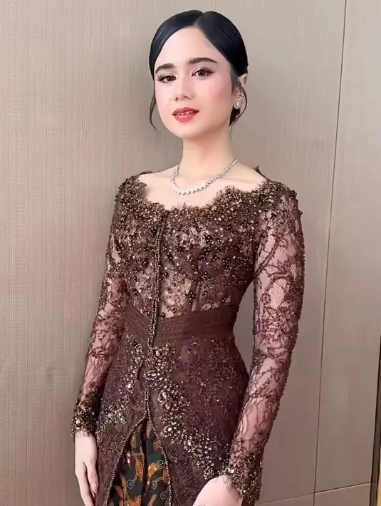 Siluet kebaya yang fitted mengikuti lekuk tubuhnya mempertegas kesan feminin, sementara aksen transparan di bagian lengan memberi sentuhan modern yang ringan. Bagian depan kebaya dibuat dengan potongan klasik lengkap dengan detail kancing yang memperkuat nuansa tradisional.