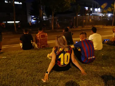 Seorang fans wanita mengenakan kaus replika nomor 10 Lionel Messi duduk di luar stadion Camp Nou di Barcelona (5/8/2021). Para fans berkumpul di depan stadion setelah Barcelona dan Lionel Messi sama-sama sepakat untuk resmi berpisah. (AP Photo/Joan Monfort)