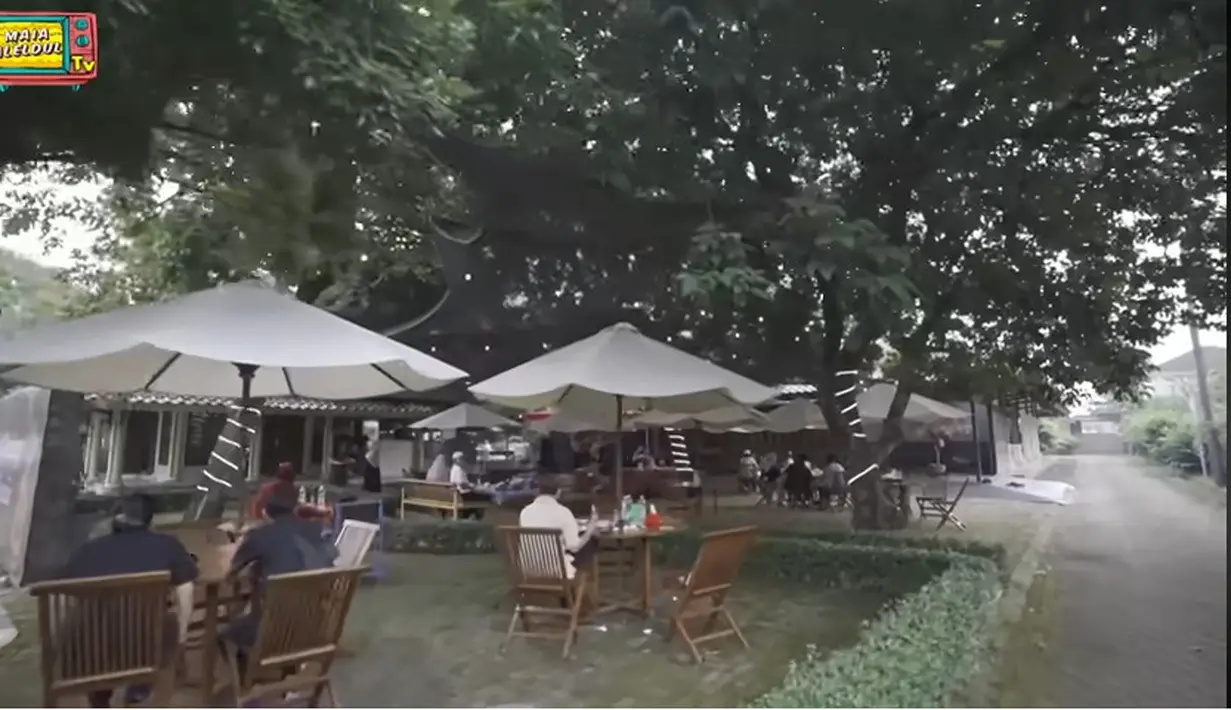 Suasana warung mandra yang diberi nama Dapur Ngaciiir. Disebelah kiri mushola terdapat spandung besar dengan tulisan selamat datang di Dapur Ngaciiir. Warung tampak nyaman di bawah pohon besar dengan beberapa payung besar. [Youtube/MAIA ALELDUL TV]