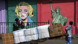 Pekerja membawa barang-barang di gerobak sambil melihat seniman melukis mural pencegahan virus corona di Mumbai (26/3/2021). India juga tengah mengalami lonjakan mengkhawatirkan yang membuat negara tersebut akan kembali melakukan pembatasan pada pertemuan publik. (AP Photo/Rajanish Kakade)