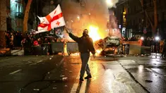 Seorang pria mengibarkan bendera nasional Georgia dekat barikade yang terbakar saat pengunjuk rasa lainnya berdiri tidak jauh dari Gedung Parlemen Georgia di Tbilisi, Georgia, Kamis (9/3/2023). Polisi menembakkan meriam air dan gas air mata untuk membubarkan demonstran di sekitar Gedung Parlemen Georgia yang memprotes rancangan undang-undang (RUU) "Agen Asing". (AP Photo/Zurab Tsertsvadze)