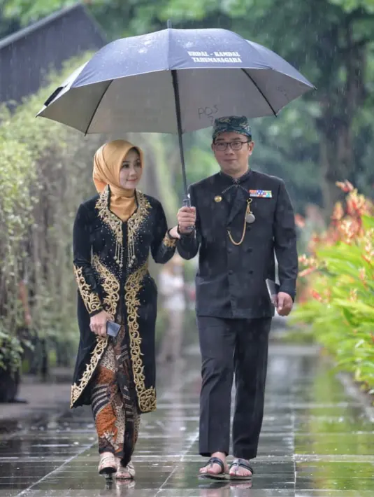 Berjalan di tengah hujan di bawah payung bersama sang suami, Atalia tampil dengan kebaya panjang yang formal. Kebaya hitam dengan aksen bordir dan payet emas ini memiliki siluet yang lebih megah daripada kebaya biasa. Atalia memadukannya dengan kain batik serasi sebagai bawahan. Foto: Instagram.