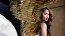 Dari samping, sayap bulu emas yang besar dan detail menjadi sorotan utama. Cinta Laura menatap kamera dengan rambut gelombang panjang dan makeup bernuansa bronze, sementara gaun putih yang melambai menyempurnakan kesan dewi dalam potret ini. [@claurakiehl]