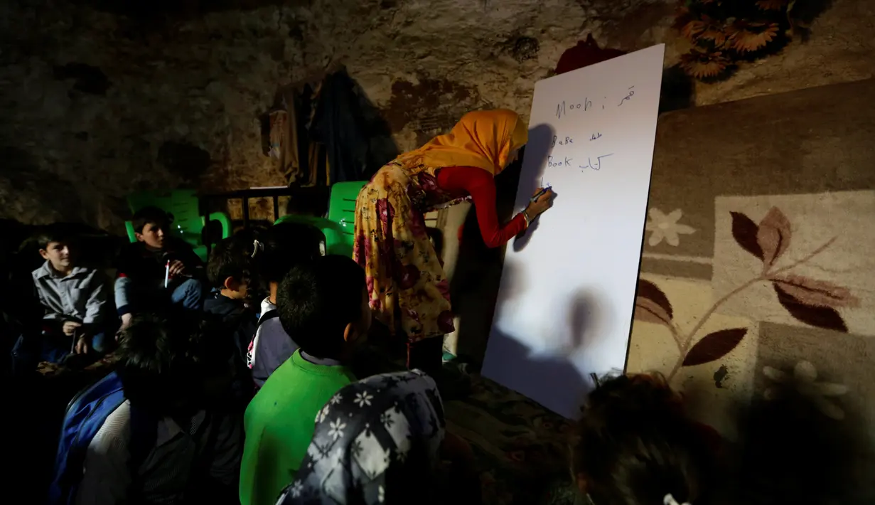 Seorang guru saat mengajarkan anak muridnya di dalam sebuah gua di desa yang dikuasai pemberontak di desa Tramla, Idlib, Suriah, 27 Maret 2016. Banyaknya anak-anak yang ingin bersekolah, kegiatan belajar dibagi menjadi dua sesi. (REUTERS/Khalil Ashawi)