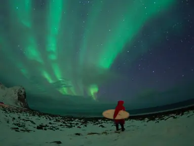 Peselancar berdiri dibawah cahaya Aurora Borealis di pantai bersalju Flackstad di Kepulauan Lofoten, Lingkar Arktik, pada 10 Maret 2016. Peselancar dari seluruh dunia datang ke Lofoten untuk berselancar di kondisi ekstrim. (AFP/Olivier Morin)