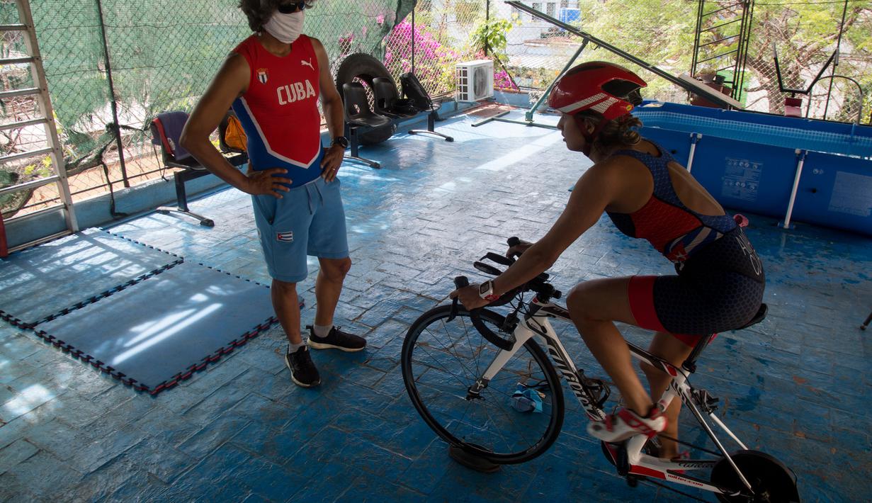 Atlet triathlon Kuba, Leslie Amat mengendarai sepedanya, diadaptasi menjadi stasioner, di bawah pengawasan pelatihnya Dioseles Fernandez di teras rumahnya di Havana, Senin (20/4/2020). Atlet Kuba tak bisa berlatih di fasilitas olahraga seperti biasanya selama pademi corona. (AP /Ismael Francisco)