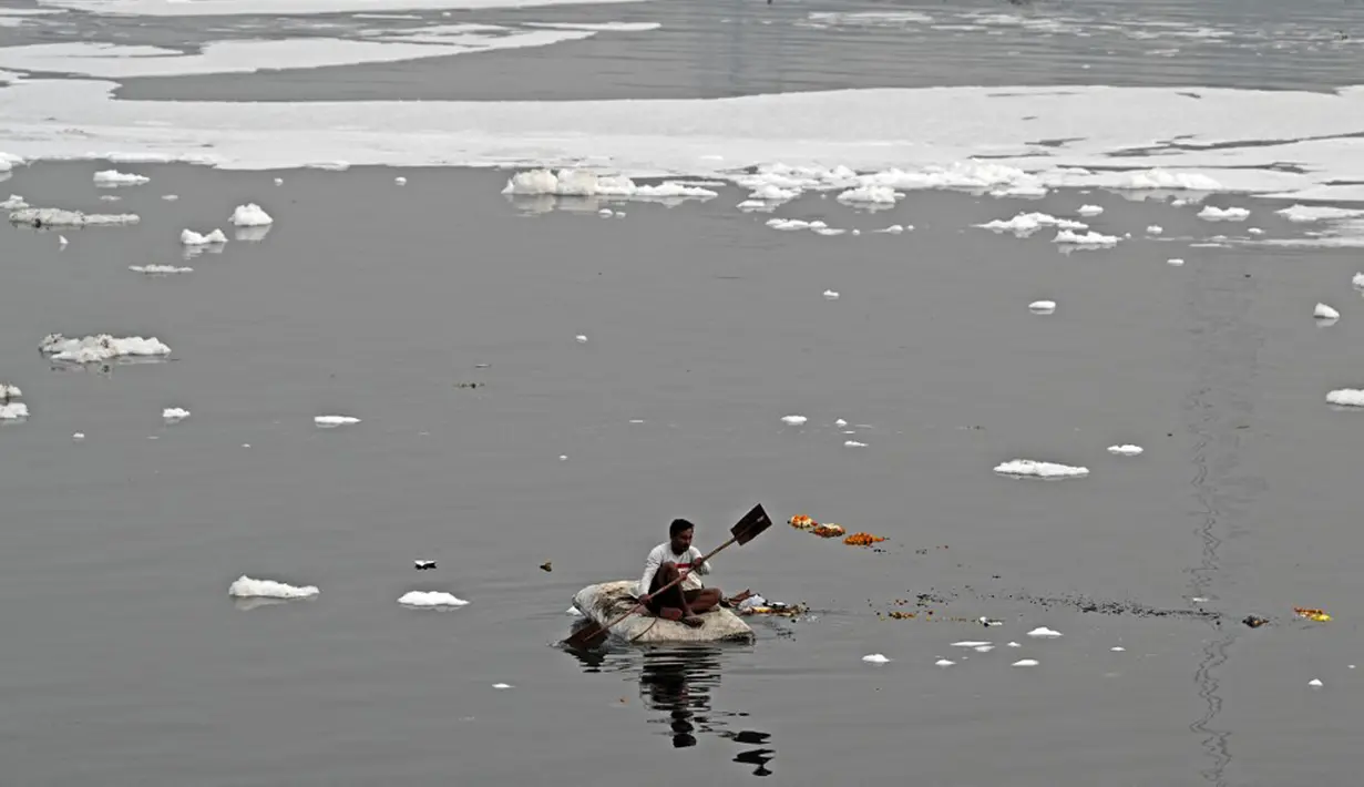Seorang pria mendayung rakitnya sambil mengumpulkan bahan-bahan daur ulang dekat potongan busa tercemar yang mengambang di Sungai Yamuna, New Delhi, India, 26 Desember 2021. Sungai Yamuna tercemari limbah beracun dari industri. (Sajjad HUSSAIN/AFP)