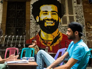 Dua pemuda duduk bersantai dengan latar belakang mural bintang Liverpool, Mohamed Salah di sebuah kedai kopi di pusat kota Kairo, 30 April 2018. Mural pemain timnas Mesir tersebut menghiasi sudut negara asalnya. (AFP Photo/KHALED DESOUKI)