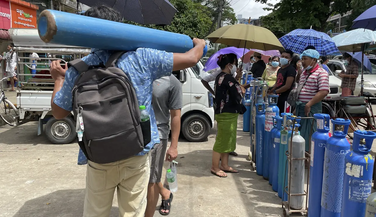 Seorang pria membawa tangki oksigen sementara yang lain berbaris untuk mengantre di luar stasiun pengisian ulang oksigen di kota Pazundaung di Yangon, Minggu (11/7/2021). Myanmar mengalami lonjakan kasus COVID-19, memicu kelangkaan pasokan oksigen yang sangat dibutuhkan pasien. (AP Photo)