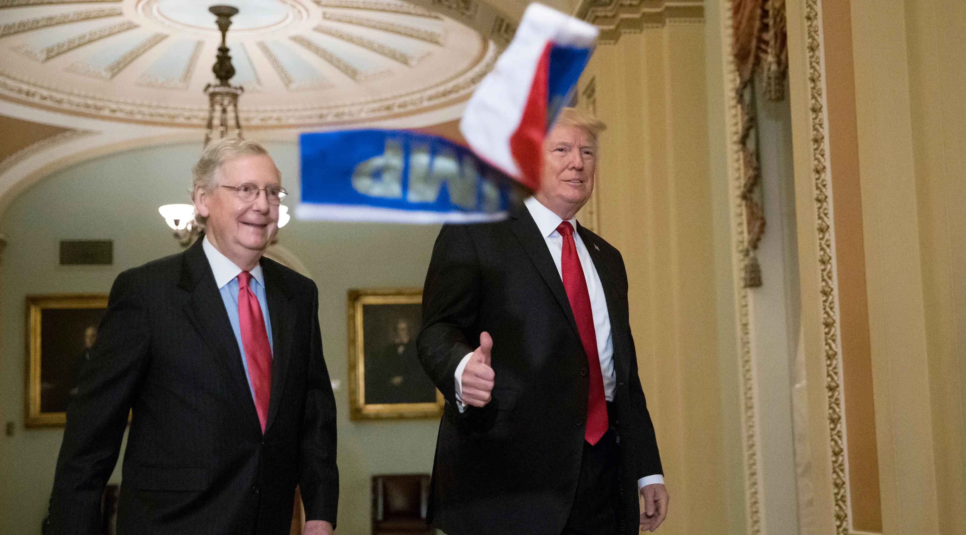 Presiden AS, Donald Trump mengacungkan jempol saat bendera Rusia berukuran kecil dilemparkan ke arahnya di gedung Capitol Hill, Washington DC, Selasa (24/10). Terlihat Trump tengah berjalan bersama senator Mitch McConnel. (AP/J. Scott Applewhite)