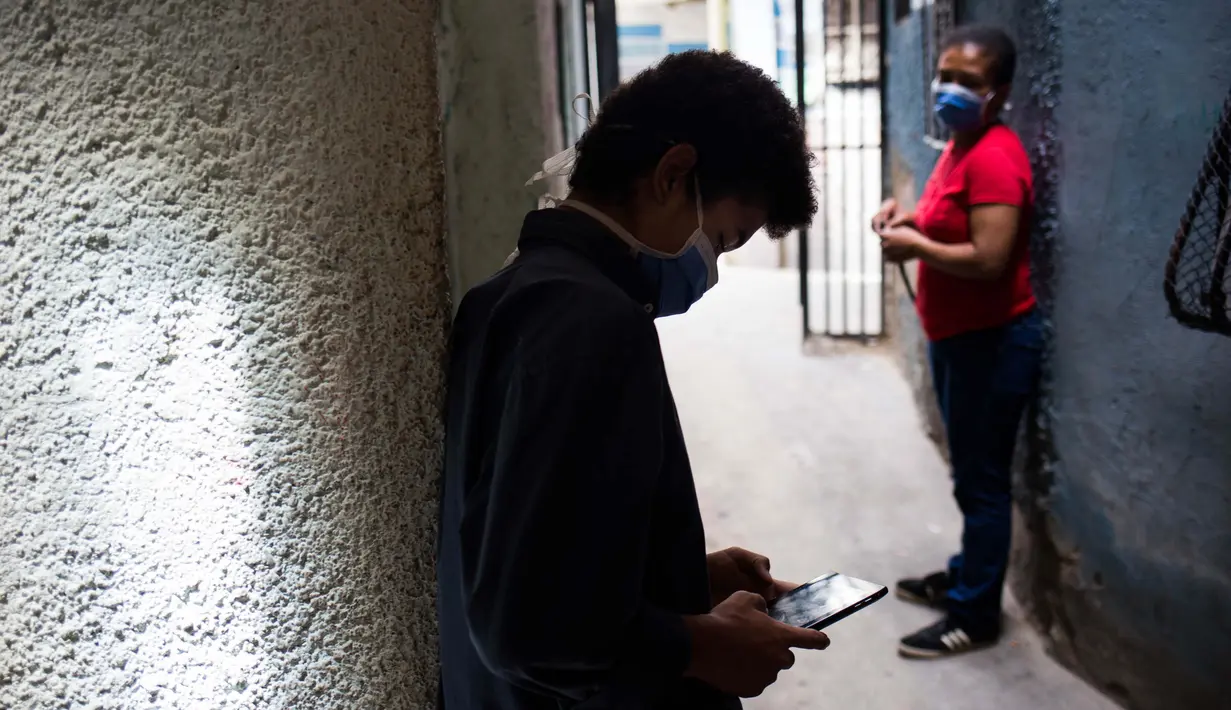 Jonathan Figueroa, 14, menggunakan ponselnya untuk terhubung ke sinyal wifi tetangga, di luar rumahnya, saat sang ibu Viviana Rodriguez mengawasinya di tengah pandemi virus corona di Bello Campo di Chacao, Caracas, Venezuela pada 6 Oktober 2020. (Cristian Hernandez/AFP)