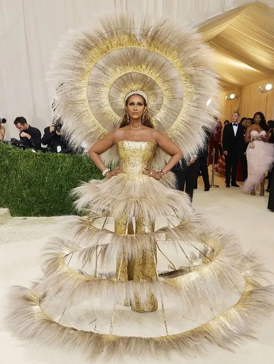 Model, artis, dan entrepreneur Iman tampil dramatis dengan jumpsuit berlapis emas dari Harris Read dengan feather tiered rings. Headpiece berbulunya pun memperkuat kesan dewi pada penampilannya. (Instagram/madalinegardener).