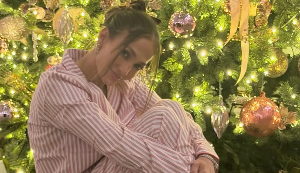 Jennifer Lopez berfoto di depan pohon Natal besar dengan ornamen yang cantik. Ia tampak mengenakan setelan piyama warna pink. [@jlo]