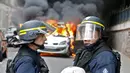 Dua petugas kepolisian berada di dekat mobil dinas yang terbakar akibat demo buru di Paris, Prancis (18/5). Demo buruh tersebut menentang kekerasan polisi dan melawan reformasi hukum yang berakhir bentrok dengan polisi. (REUTERS / Charles Platiau)