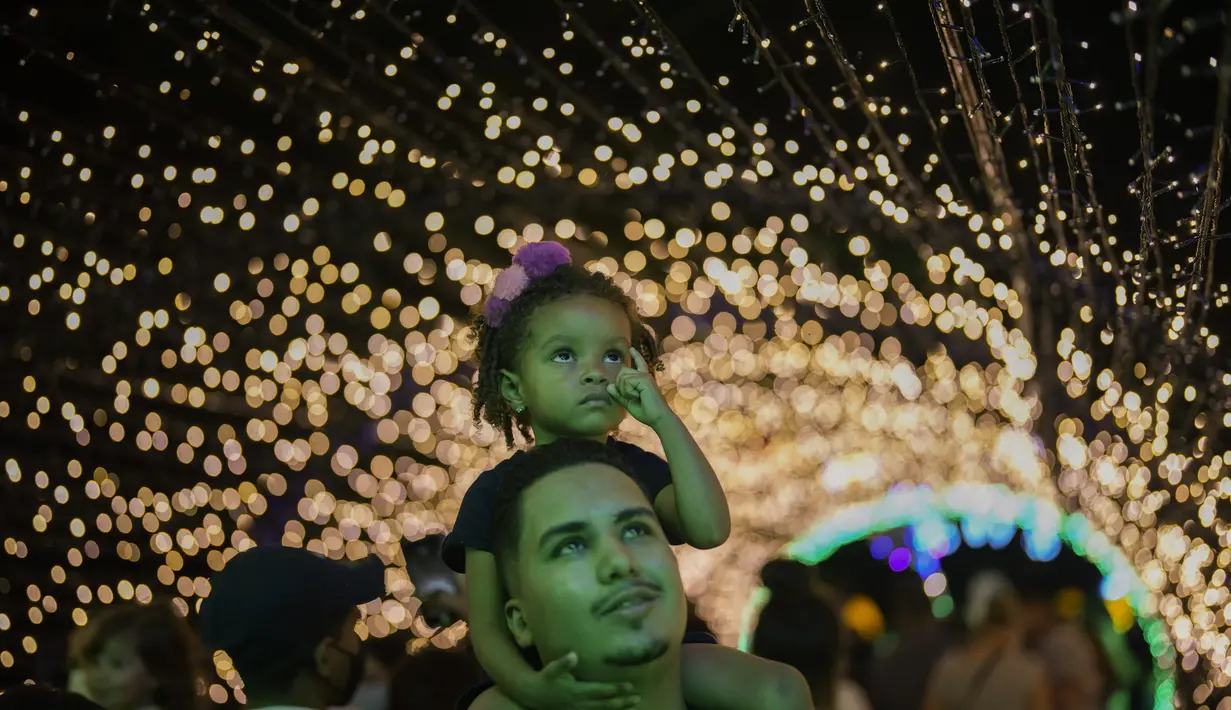 Seorang pria menggendong putrinya saat acara lampu Natal di taman Ibirapuera di Sao Paulo, Brasil, Selasa (14/12/2021). Jelang perayaan Natal, taman dan pantai dihiasi lampu-lampu yang indah. (AP Photo/Andre Penner)