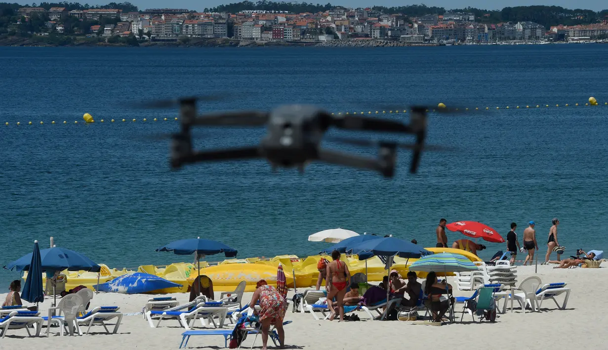 Sebuah drone atau pesawat tak berawak yang diterbangkan oleh polisi setempat memperingatkan orang-orang untuk menjaga jarak di pantai Areas di Sanxenxo, Spanyol, Selasa (14/7/2020). Drone tersebut untuk memantau jaga jarak antarwisatawan saat berlibur di masa pandemi Corona. (MIGUEL RIOPA / AFP)