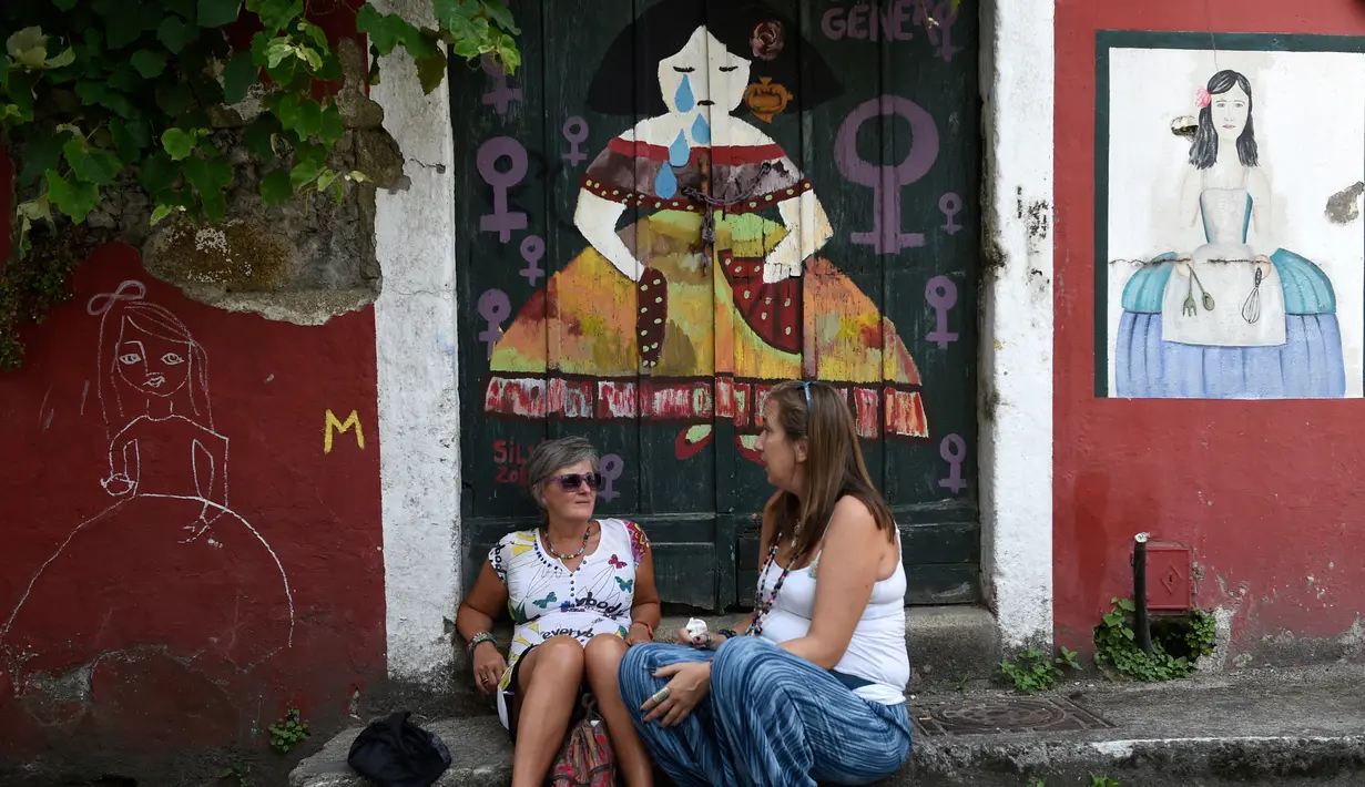 Dua wanita duduk di samping mural karya 'Meninas' selama festival seni jalanan tahunan Meninas de Canido di Ferrol, Spanyol, Minggu (2/9). (MIGUEL RIOPA/AFP)