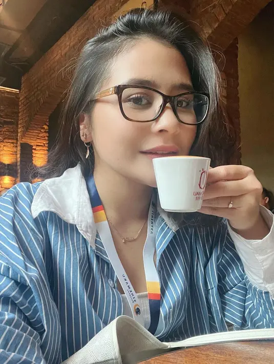 Potret selfie Prilly minum kopi mengenakan kemeja lengan panjang berwarna biru bermotif garis dengan riasan wajah minimalis dan kacamata. [Foto: Instagram/prillylatuconsina96]