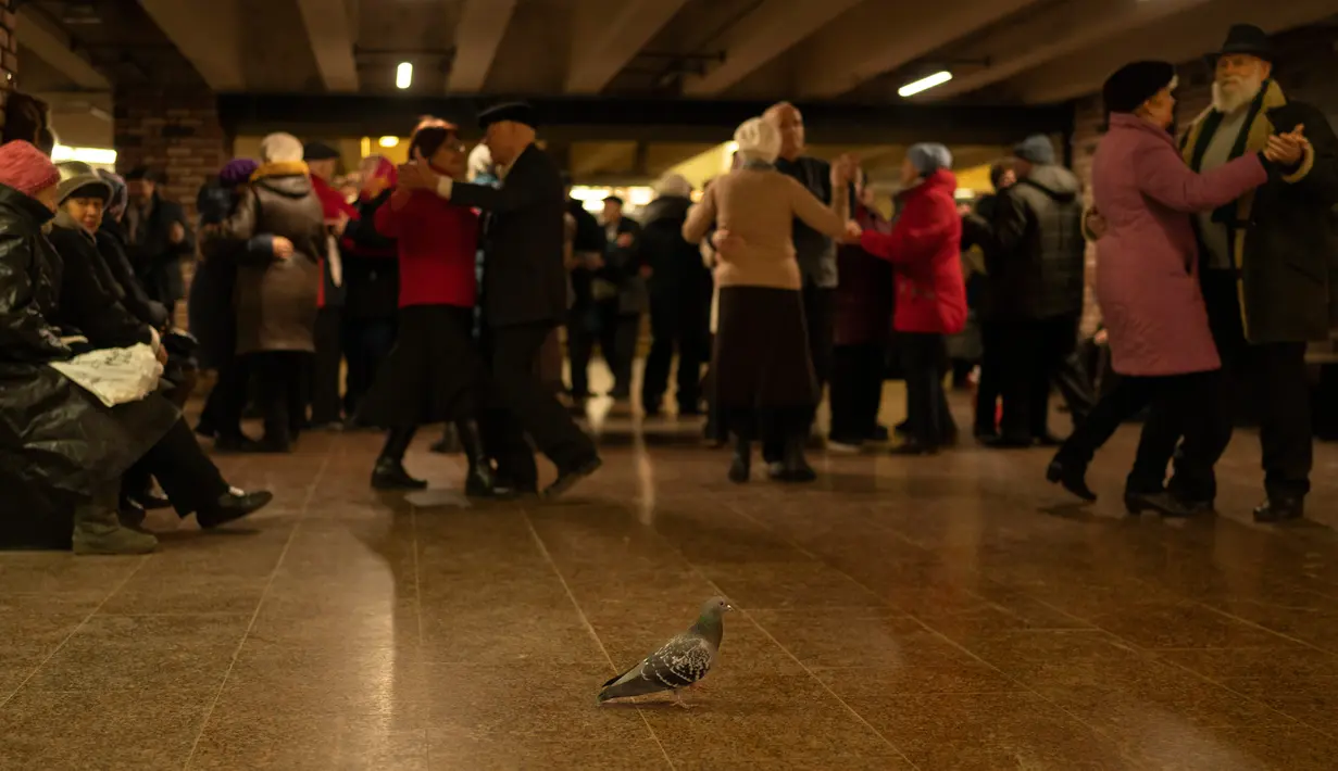 Seekor merpati berjalan dilatarbelakangi oleh pasangan lansia yang sedang menari di Stasiun Kereta Bawah Tanah Teatralna, Kiev, Ukraina, 5 Maret 2023. Puluhan pensiunan menari selama lebih dari dua jam dalam acara akhir pekan yang telah berlangsung selama puluhan tahun dan hanya sempat terganggu oleh invasi Rusia ke Ukraina. (AP Photo/Vadim Ghirda)