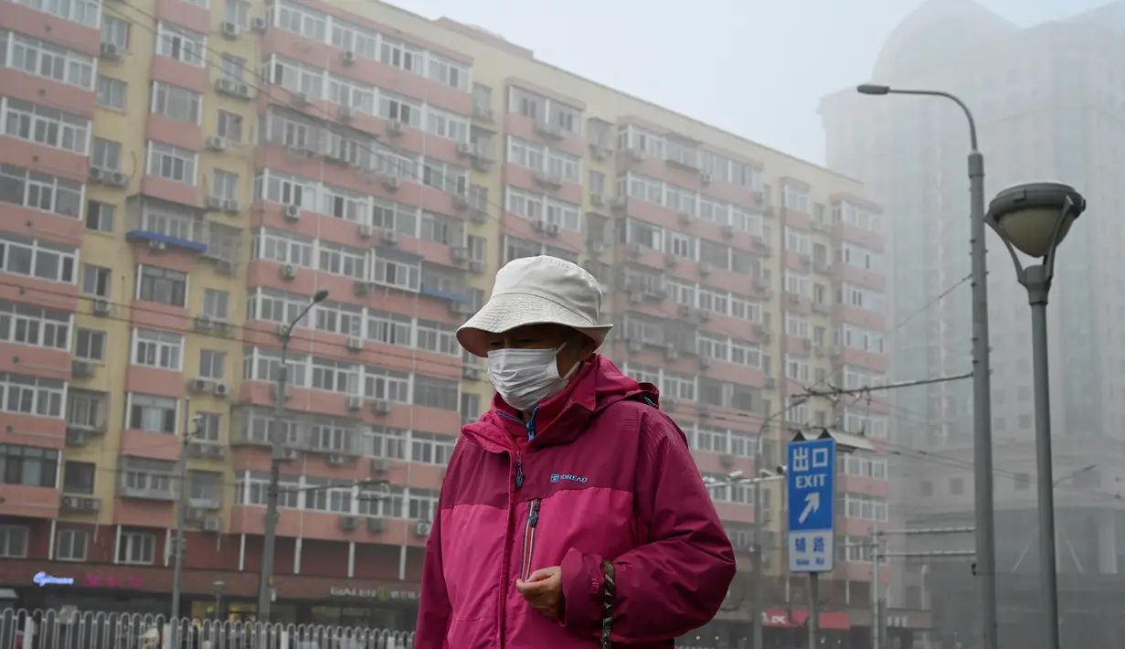 Seorang perempuan yang mengenakan masker berjalan di sepanjang jalan yang berkabut dan tercemar di Beijing, Sabtu (6/11/2021). Jalan raya dan taman bermain sekolah di Beijing ditutup pada Jumat (5/11) karena polusi kabut asap yang tebal, setelah China meningkatkan produksi batu bara. (JADE GAO/AFP)