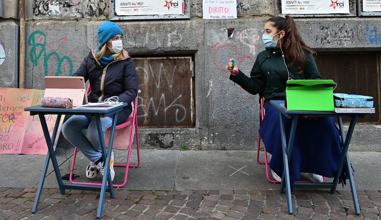 Anita Iacovelli (kiri) dan temannya Lisa Rogliatti, berusia 12 tahun, duduk di depan sekolah Italo Calvino di Turin, Italia, 17 November 2020. Anita memprotes penutupan sekolahnya karena pembatasan COVID-19 dengan duduk di luar gedung sambil mengikuti pelajaran jarak jauh. (Miguel MEDINA/AFP)