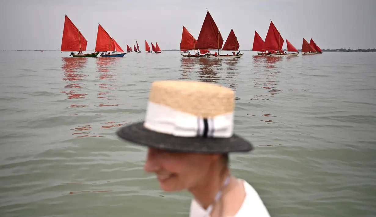 Seorang wanita berdiri di depan anggota asosiasi pelayaran budaya yang berlayar dalam acara Regatta Merah di Venesia, Italia, Minggu (20/6/2021). Red Regatta mengumpulkan lebih dari 50 perahu tradisional Venesia dengan 52 layar warna merah. (MARCO BERTORELLO/AFP)