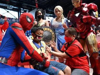 Dua orang berkostum superhero berpose dengan seorang anak yang menerima vaksin Covid-19 Pfizer-BioNtech untuk anak-anak berusia 5-11 di sebuah gym di San Juan City, pinggiran kota Manila, Filipina pada 7 Februari 2022. (Ted ALJIBE / AFP)