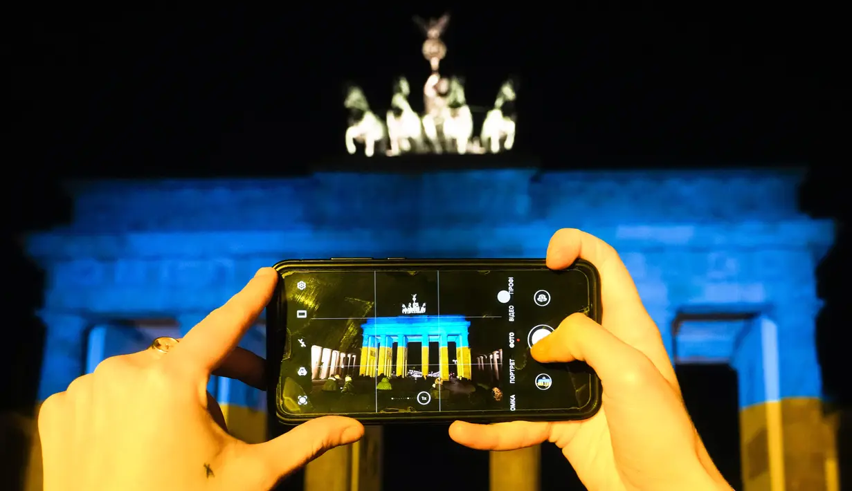 Seorang perempun mengambil foto Gerbang Brandenburg setelah diterangi dengan warna bendera Ukraina di Berlin, Jerman, Rabu (23/2/2022). Salah satu bangunan ikonik itu diterangi warna biru dan kuning untuk menunjukkan solidaritas selama ketegangan Ukraina dengan Rusia. (AP Photo/Markus Schreiber)