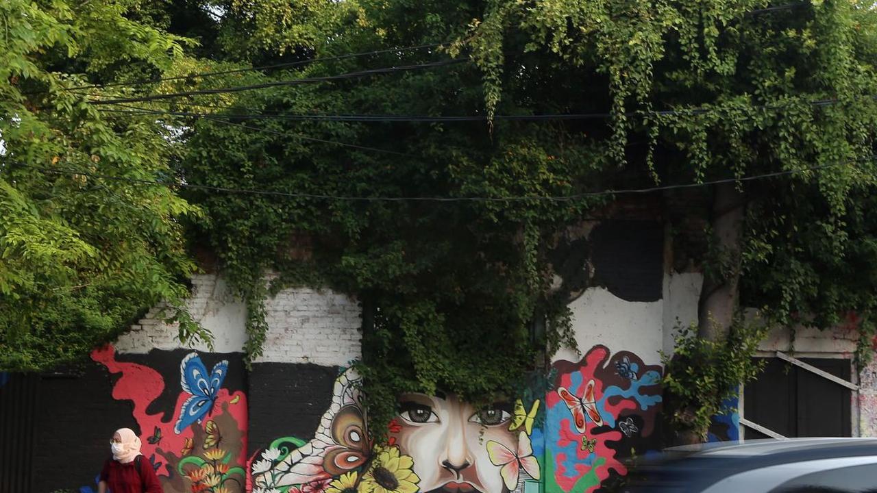 Seorang warga tampak sedang menunggu kendaraan umum di trotoar dengan latar belakang pemandangan mural karya petugas PPSU di kawasan Jalan Malaka, Jakarta.