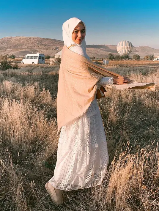 Anggunnya Citra Kirana mengenakan hijab dan dress panjang berwarna putih dipadu dengan syal krem dengan latar belakang balon udara dan padang ilalang di Cappadocia, Turki. (Liputan6.com/IG/@citraciki)