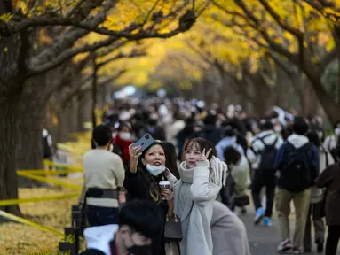 Dua wanita yang memakai masker  untuk membantu mengekang penyebaran virus corona berselfie saat mereka berjalan melewati deretan pohon ginkgo di sepanjang trotoar saat pepohonan dan trotoar ditutupi dengan daun kuning cerah di Tokyo (29/11/2021). (AP Phot/Kiichiro Sato)